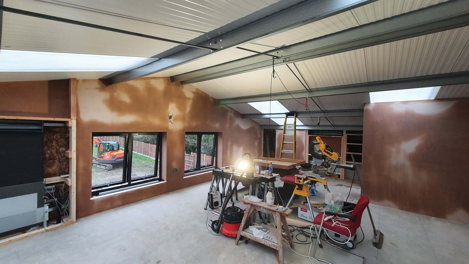 Large room under construction with steel beams and roof lights, walls freshly plastered, tools on trestles